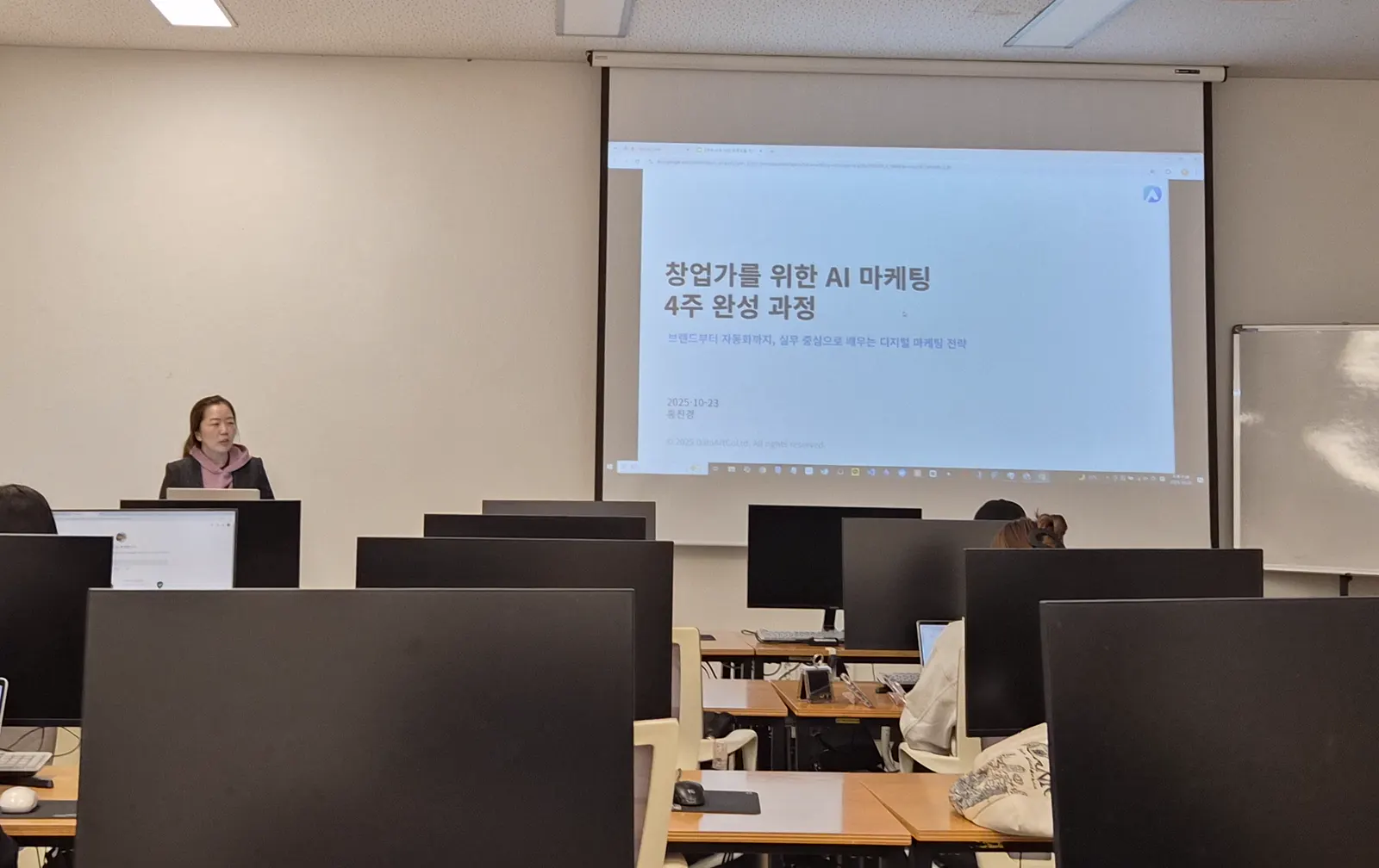 컴퓨터 실습실 앞쪽 스크린에 창업가를 위한 AI 마케팅 과정 첫 화면이 떠 있고, 강사가 교탁에서 설명하는 모습