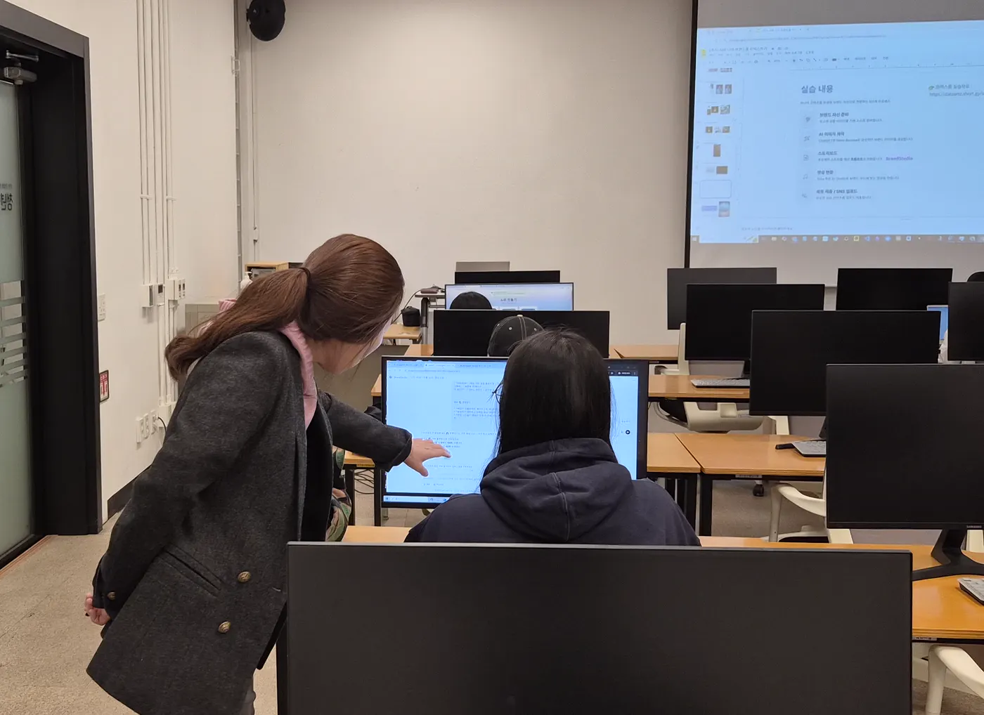 강사가 수강생 옆에서 모니터 화면을 가리키며 실습을 안내하는 뒷모습