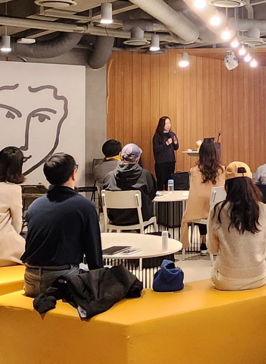 참석자들이 뒷모습으로 앉아 있고, 앞에서 강사가 마이크를 들고 설명하는 모습