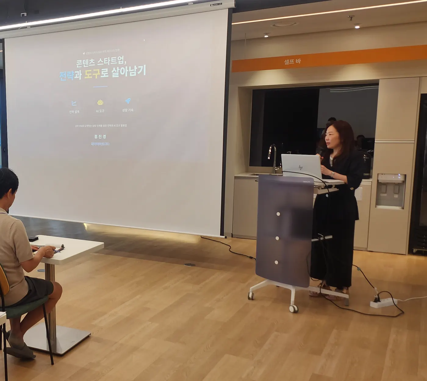 스크린에 콘텐츠 스타트업 강연 제목이 떠 있고 강사가 연단에서 발표하는 모습